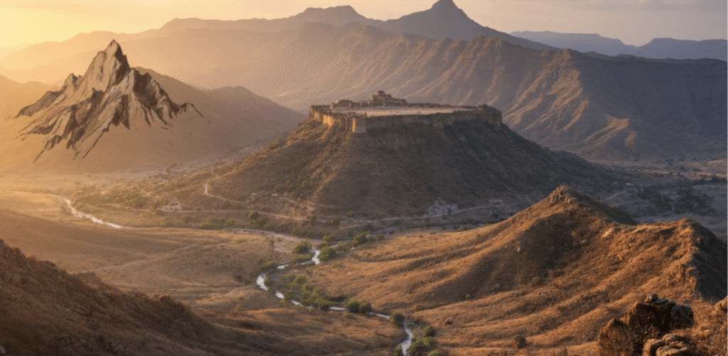 A scenic landscape of the Aravalli mountain range in Rajasthan featuring rugged peaks, flat-topped plateaus, and dry valley vegetation under a sunset sky.